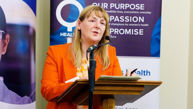 Blonde lady in orange blazer giving speech infront of two vertical banners
